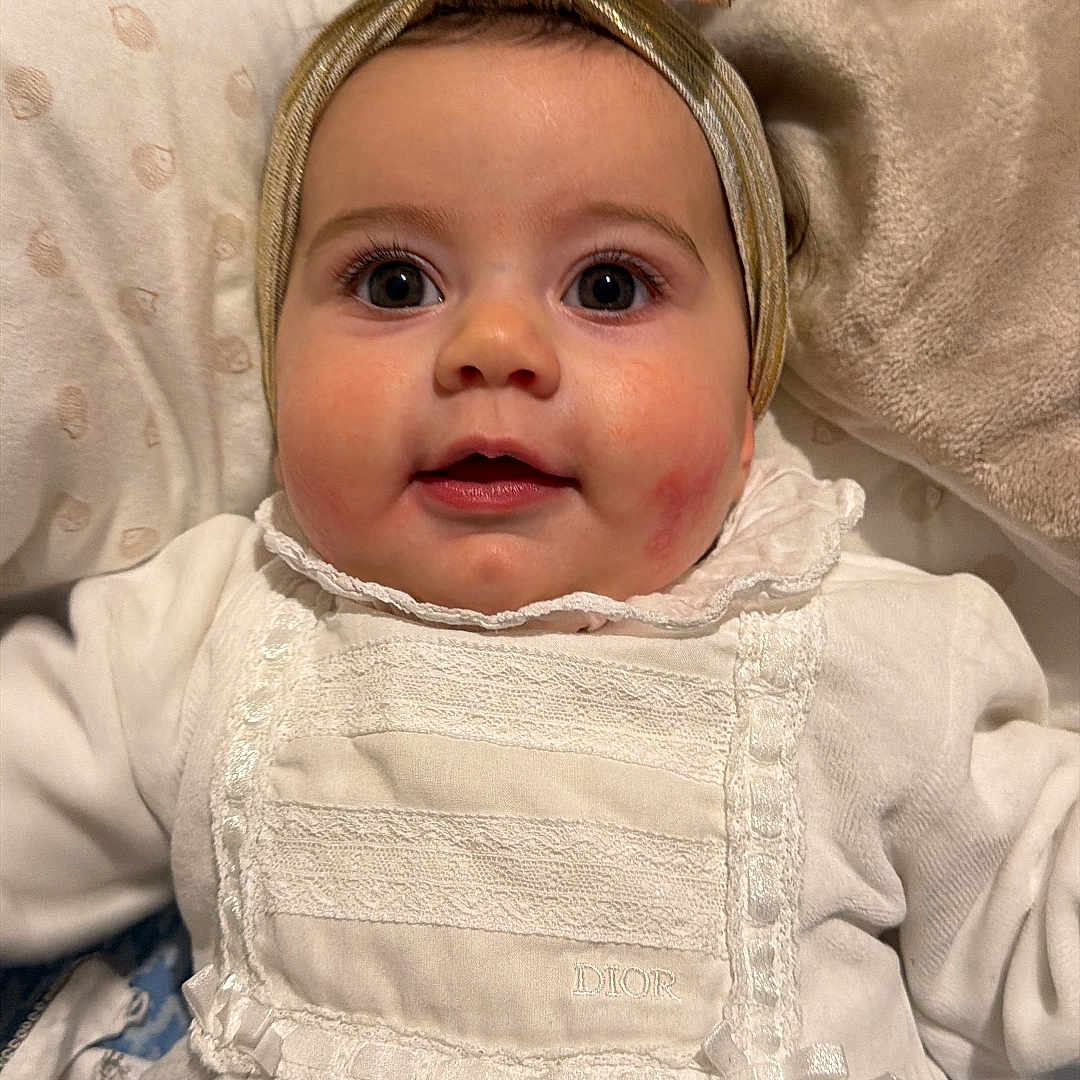 Chiara participe au concours pour gagner de l'argent avec cette photo : baby, bedding, big_eyes, blanket, bow, cheeks, child, close_up, face, headband, indoor, infant, lace, lying_down, onesie, pillow, portrait, smile, soft_lighting, white_clothing