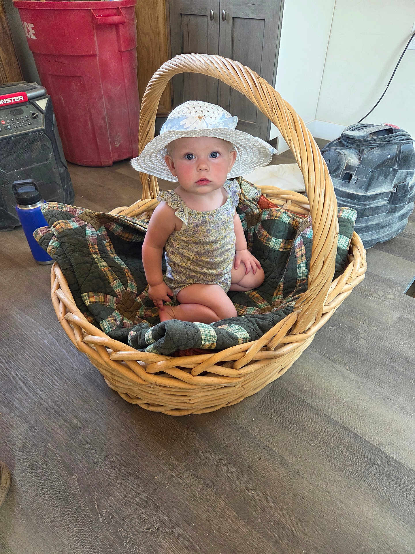 Jane joined the competition — help win amazing prizes! baby, basket, hat, quilt, indoor, wood_floor, child, wicker, curious, blue_eyes, sitting, plaid, blanket, face, person, floor, cabinet, red_bin, tool, expression