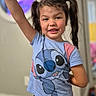 child, girl, smiling, portrait, indoors, standing, raised_arm, pigtails, hair_bows, blue_shirt, stitch_character, black_shorts, wood_floor, home, playful, happy, face, person, casual_clothing, colorful_background