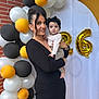 woman, baby, balloons, black_dress, headband, portrait, celebration, party, gold_balloons, white_backdrop, brick_wall, smile, holding, person, fashion, event, happy, cute, indoor, family