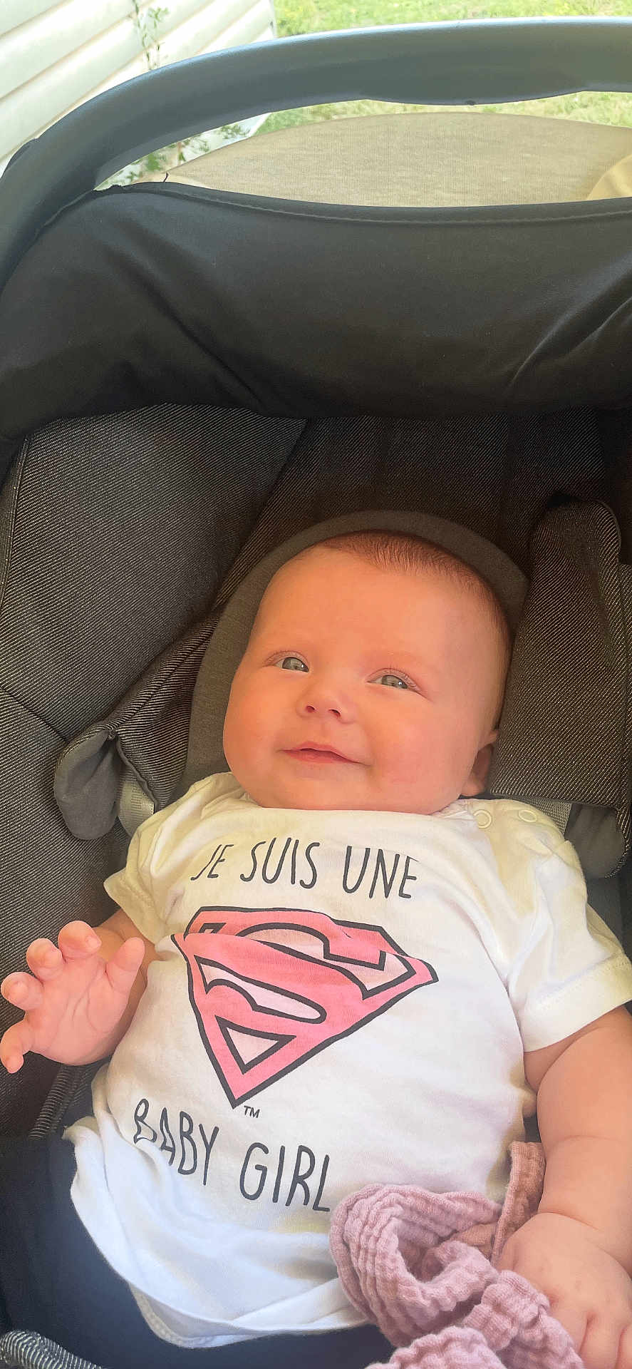 Eden a rejoint le concours — aidez-le/la à gagner de superbes lots ! baby, infant, smiling, car_seat, white_shirt, superman_logo, pink, clothing, child, young_child, cute, portrait, hand, fabric, seat, outdoor, greenery, comfort, happy, toddler
