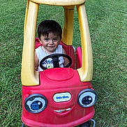 Fred is registered to the contest to win money with this photo: toddler, toy_car, grass, outdoor, child, smiling, steering_wheel, plastic, red, yellow, people, chairs, gathering, sunny, lawn, happy, casual, clothing, play, fun