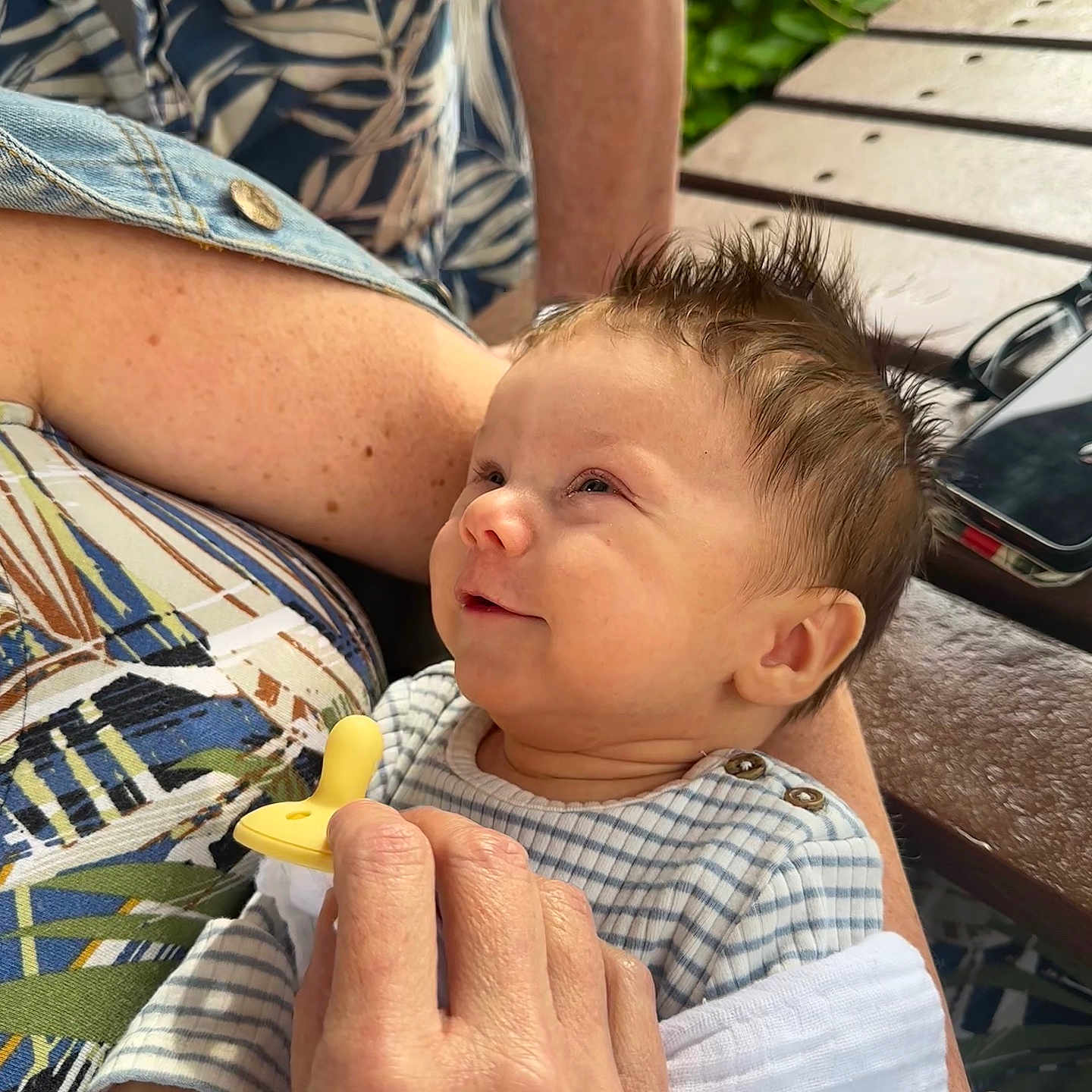 Brooks is registered to the contest to win money with this photo: arm, baby, child, closeup, clothing, face, greenery, hair, hand, holding, infant, outdoor, pacifier, person, skin, smile, striped, table, watch, wood