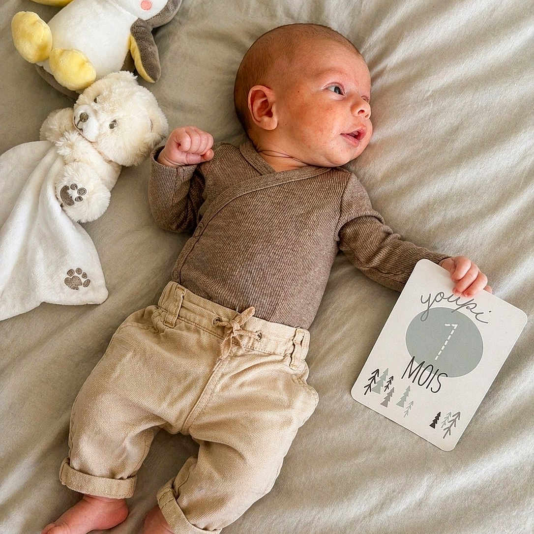 Tyméo participe au concours pour gagner de l'argent avec cette photo : baby, infant, plush_toy, penguin_toy, teddy_bear, milestone_card, 7_months, bed, blanket, clothing, pants, long_sleeve, bare_feet, cute, lying_down, portrait, indoor, soft_lighting, child, smile