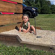 Logan is registered to the contest to win money with this photo: toddler, child, sandbox, gravel, wooden_structure, cap, outdoor, grass, truck, trees, sky, clouds, sunlight, greenery, summer, play, serious_expression, nature, casual_clothing, daytime