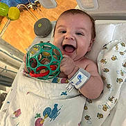 Ashton joined the competition — help win amazing prizes! baby, infant, smiling, toy, blanket, crib, hospital, wristband, playful, happy, indoor, child, person, face, hand, colorful, patterned_blanket, wooden_floor, plastic_toy, wrapped