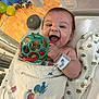 baby, infant, smiling, toy, blanket, crib, hospital, wristband, playful, happy, indoor, child, person, face, hand, colorful, patterned_blanket, wooden_floor, plastic_toy, wrapped