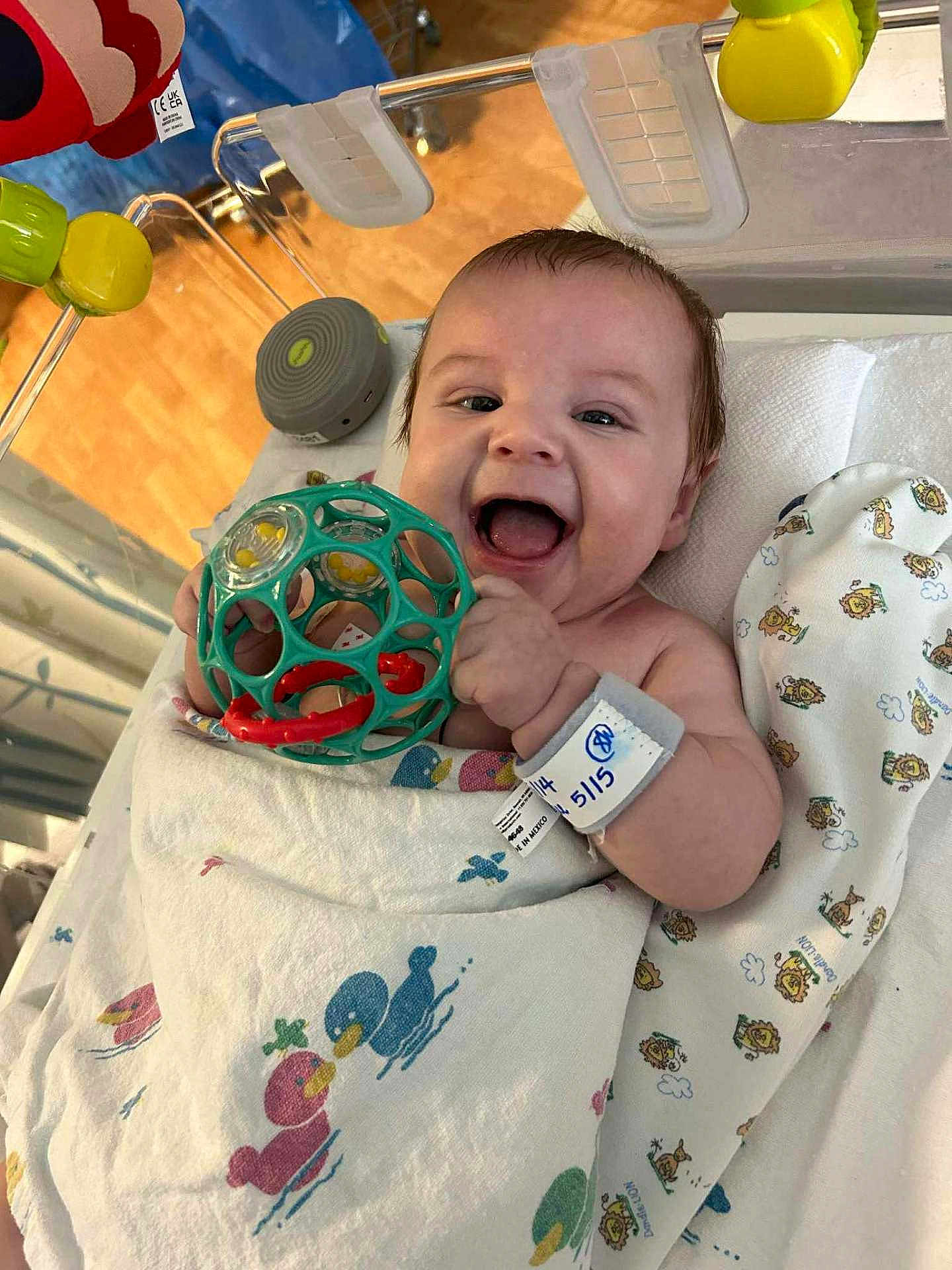 Ashton joined the competition — help win amazing prizes! baby, infant, smiling, toy, blanket, crib, hospital, wristband, playful, happy, indoor, child, person, face, hand, colorful, patterned_blanket, wooden_floor, plastic_toy, wrapped