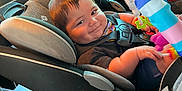 Gabriel joined the competition — help win amazing prizes! baby, car_seat, child, toy, colorful, smile, face, seatbelt, interior, car, window, sunlight, clothing, hand, seat, head, infant, sitting, cute, play