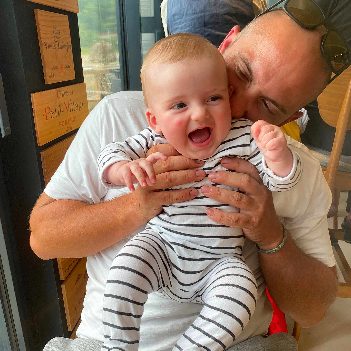 Jaime a rejoint le concours — aidez-le/la à gagner de superbes lots ! baby, casual_clothing, child, family, glasses, hug, indoor, interaction, joy, laughter, man, parent, person, playful, seated, smile, striped_onesie, table, window, wooden_decor