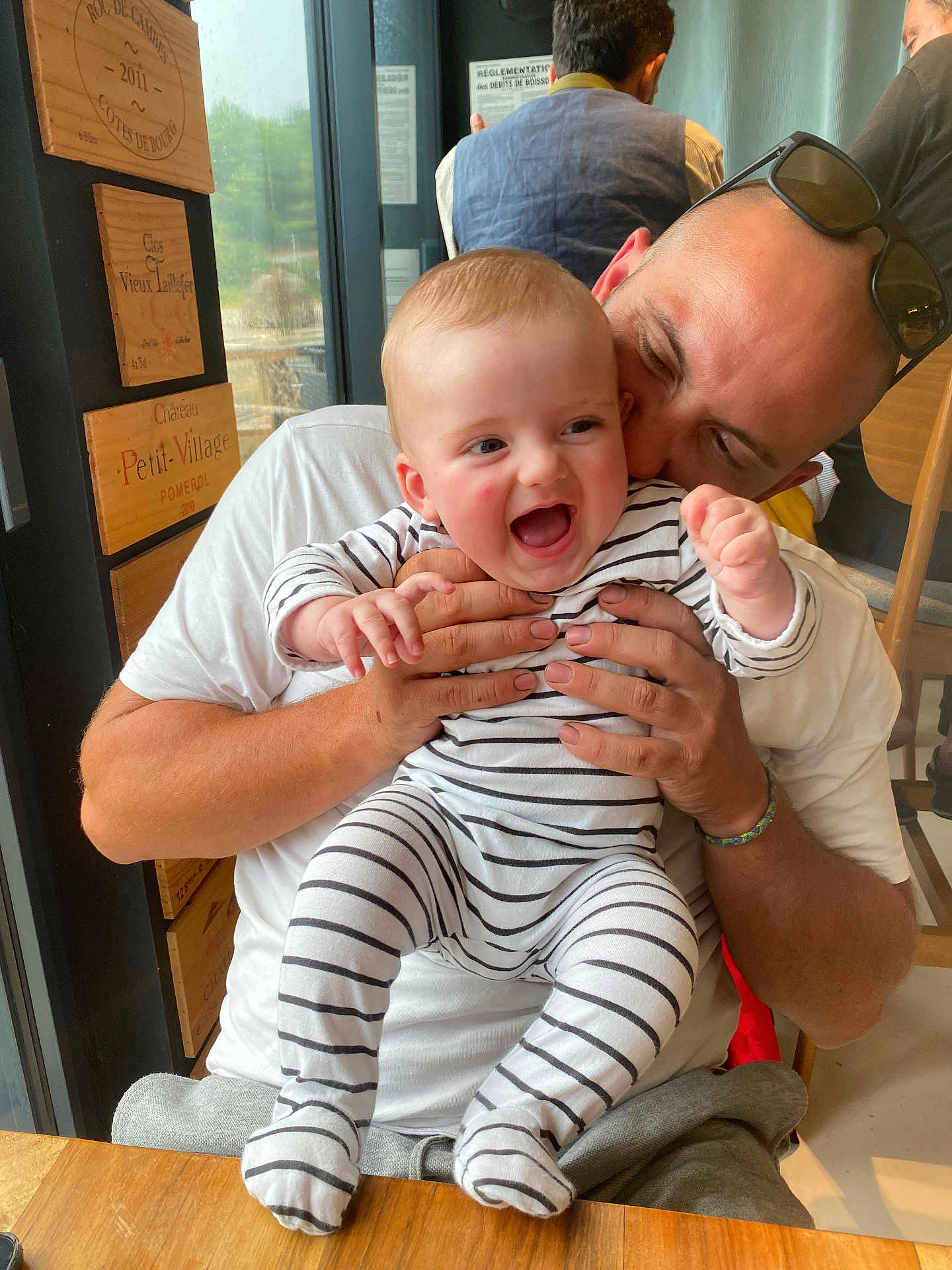 Jaime a rejoint le concours — aidez-le/la à gagner de superbes lots ! baby, man, striped_onesie, indoor, hug, laughter, joy, playful, glasses, table, wooden_decor, window, person, child, casual_clothing, smile, family, parent, interaction, seated