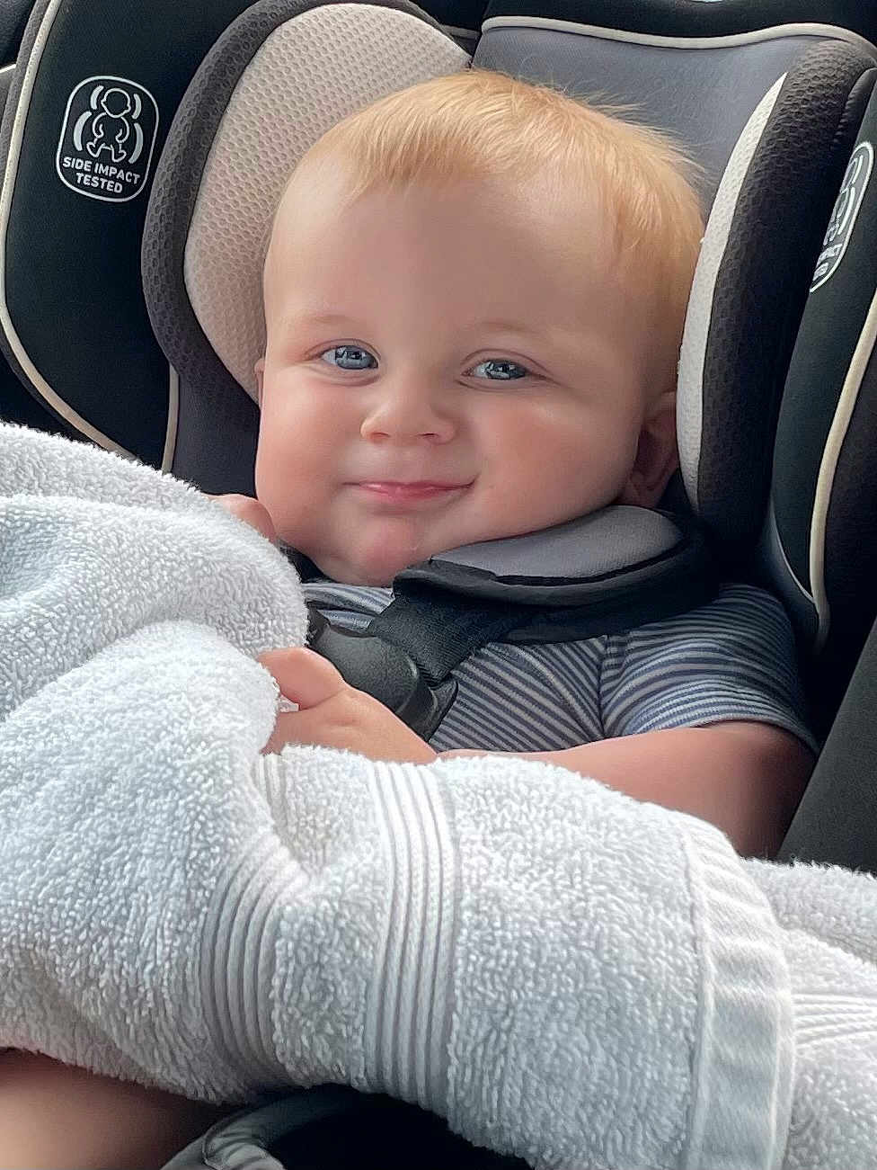 Bryson is registered to the contest to win money with this photo: baby, child, car_seat, seatbelt, blue_eyes, blond_hair, towel, smile, cute, infant, striped_clothing, safety, indoors, closeup, face, headrest, comfort, car_interior, soft_texture, happy