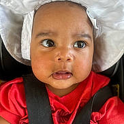Alora joined the competition — help win amazing prizes! baby, child, face, hat, white_hat, red_shirt, car_seat, black_straps, wide_eyes, curious, close_up, infant, skin, young_child, portrait, seatbelt, cute, expression, headwear, indoors
