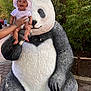 Alora joined the competition — help win amazing prizes! baby, child, panda_statue, outdoor, greenery, hat, smiling, person, hand, nature, tree, statue, footpath, sunlight, park, casual_clothing, happy, playful, daytime, people_in_background