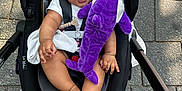 Alora is registered to the contest to win money with this photo: baby, child, car_seat, stroller, plush_toy, purple_toy, outdoor, pavement, feet, sandals, person, clothing, happy, cute, infant, seatbelt, black_car_seat, skin, hand, foot