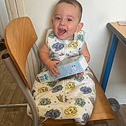 Toni participe au concours pour gagner de l'argent avec cette photo : toddler, child, smiling, chair, wooden_chair, onesie, cartoon_print, indoor, floor, box, happy, person, baby, clothing, furniture, flooring, table, wall, head, sitting
