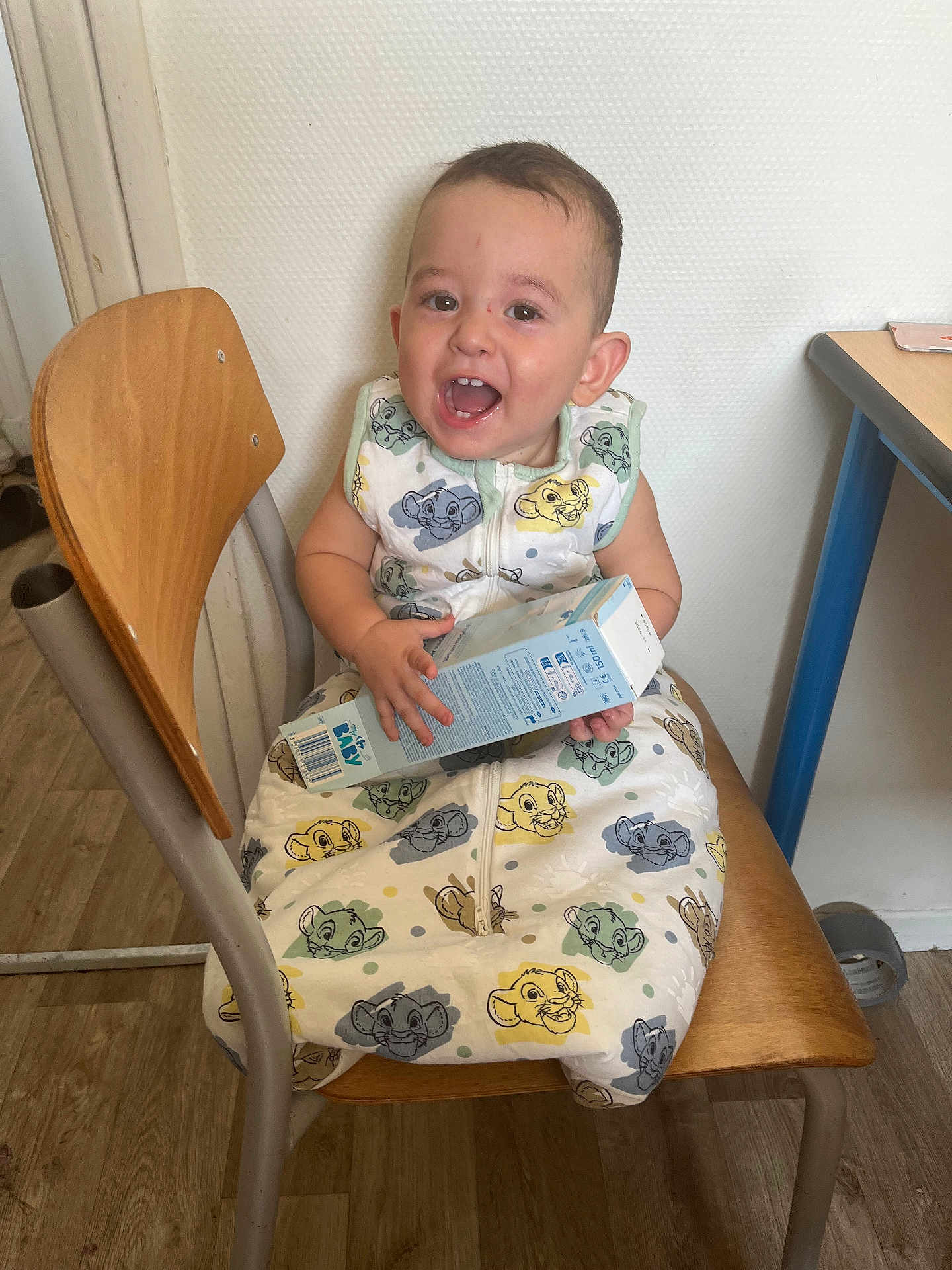 Toni participe au concours pour gagner de l'argent avec cette photo : toddler, child, smiling, chair, wooden_chair, onesie, cartoon_print, indoor, floor, box, happy, person, baby, clothing, furniture, flooring, table, wall, head, sitting