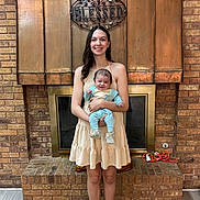 Zach is registered to the contest to win money with this photo: woman, baby, child, smiling, holding, dress, fireplace, brick_wall, indoor, home, decor, sign, blessed, floor_rug, footwear, happy, portrait, person, family, cozy