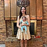woman, baby, child, smiling, holding, dress, fireplace, brick_wall, indoor, home, decor, sign, blessed, floor_rug, footwear, happy, portrait, person, family, cozy