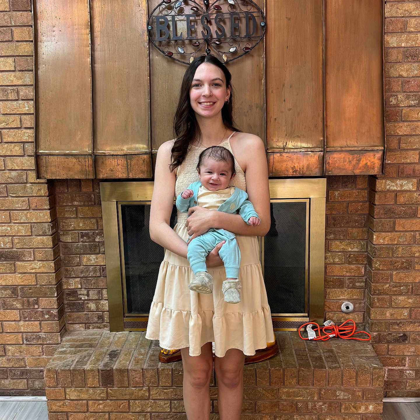 Zach is registered to the contest to win money with this photo: woman, baby, child, smiling, holding, dress, fireplace, brick_wall, indoor, home, decor, sign, blessed, floor_rug, footwear, happy, portrait, person, family, cozy