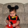 toddler, child, costume, mickey_mouse, mouse_ears, yellow_bow_tie, red_overalls, black_shirt, remote_control, couch, furniture, indoors, cute, baby, chubby, hands, sitting, focused, face, hair