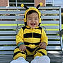 Liam joined the competition — help win amazing prizes! baby, child, costume, bee_costume, smiling, bench, outdoor, happy, cute, footwear, white_shoes, bracelet, person, portrait, infant, yellow, black, headgear, playful, sitting