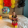 baby, smiling, birthday_cake, mickey_mouse, balloons, red, yellow, black, confetti, bed, white_bedspread, headboard, nike_box, disney_characters, first_birthday, suspenders, bowtie, celebration, indoor, happy
