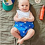 baby, infant, smiling, blue_outfit, whale_print, white_top, cap, hat, sunscreen, orange_bottle, wooden_plaque, blanket, soft_texture, lying_down, cute, happy, child, portrait, indoor, clothing
