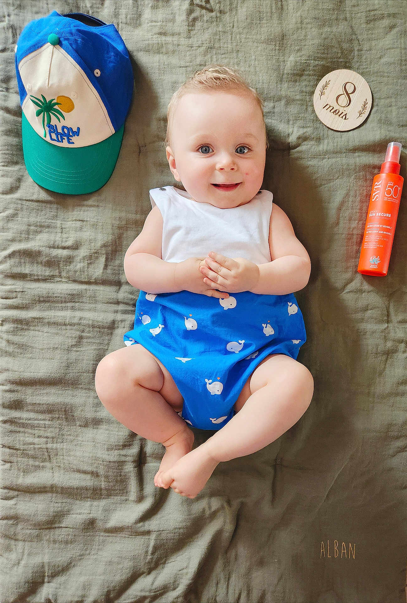 Alban participe au concours pour gagner de l'argent avec cette photo : baby, infant, smiling, blue_outfit, whale_print, white_top, cap, hat, sunscreen, orange_bottle, wooden_plaque, blanket, soft_texture, lying_down, cute, happy, child, portrait, indoor, clothing