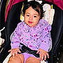 baby, child, car_seat, purple_clothing, bow, infant, cute, smiling, sitting, black_hair, indoors, seatbelt, plush_padding, face, hands, legs, skin, comfort, child_seat, young