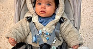 Swann participe au concours pour gagner de l'argent avec cette photo : beige_color_palette, blanket, blue_eyes, child, cozy, curious_expression, curly_hair, hands, hood, indoor, infant, pacifier, plush_toy, portrait, safety_straps, sitting, stroller, tiled_floor, toy_clip, winter_coat
