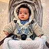 beige_color_palette, blanket, blue_eyes, child, cozy, curious_expression, curly_hair, hands, hood, indoor, infant, pacifier, plush_toy, portrait, safety_straps, sitting, stroller, tiled_floor, toy_clip, winter_coat