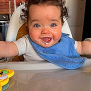 Swann participe au concours pour gagner de l'argent avec cette photo : baby, bib, blue_eyes, cheeks, closeup, curly_hair, drooling, face, feeding, happy, high_chair, infant, kitchen, portrait, reflection, refrigerator, smile, sticker, table, toy