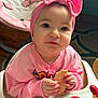 baby, child, pink_bow, headband, high_chair, bread, food, sweater, indoor, smile, face, hands, curious, seat, toy, floor_rug, cute, person, young_child, feeding