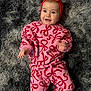 baby, child, smiling, headband, bow, pink_clothing, red_clothing, patterned_clothing, lying_down, rug, fluffy_rug, happy, cute, infant, person, portrait, indoors, soft_texture, cozy, bright_colors