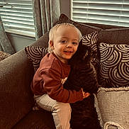 Jaxson joined the competition — help win amazing prizes! toddler, child, cat, couch, cushion, smile, indoor, window_blinds, curtains, sweatshirt, light_pants, barefoot, tabby_cat, hugging, happy, cozy, furniture, home, pet, portrait