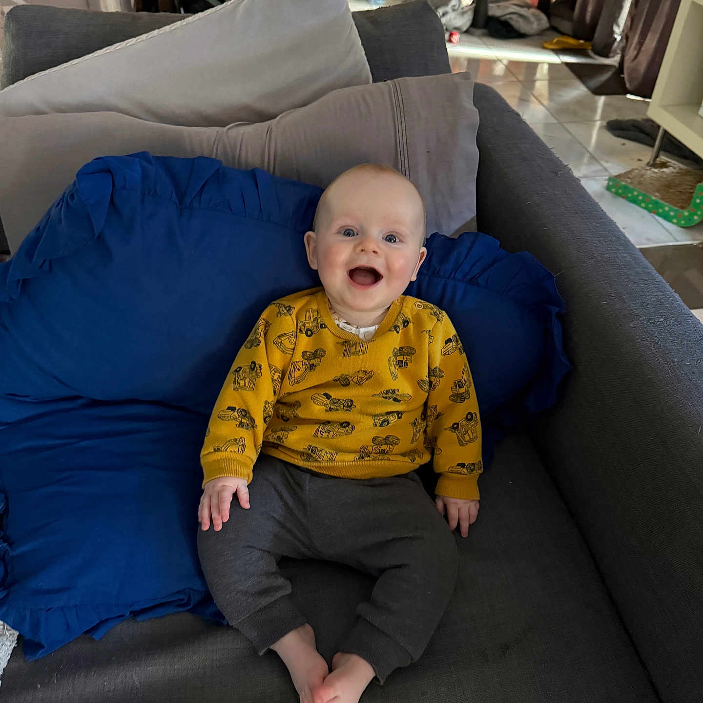 Rían is registered to the contest to win money with this photo: baby, child, smiling, sitting, couch, pillow, yellow_sweater, gray_pants, indoors, living_room, happy, face, person, barefoot, furniture, home, blue_pillow, bright_eyes, casual_clothing, cozy