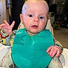 Rían joined the competition — help win amazing prizes! baby, child, high_chair, bib, teal_bib, colorful_clothing, long_sleeve, hand_gesture, curious, indoor, face, blue_eyes, blond_hair, feeding_time, kitchen, plate, seat, person, expression, young_child