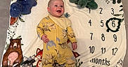 Rían is registered to the contest to win money with this photo: baby, infant, smile, yellow_onesie, dinosaur_print, plush_toys, blanket, animal_print, milestone_blanket, months_marker, happy, cute, lying_down, child, soft_toys, floor, indoors, portrait, playful, bright