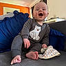 Rían joined the competition — help win amazing prizes! baby, barefoot, bear_design, blanket, blue_cushion, child, cozy, curtains, cushion, cute, grey_clothing, happy, home, indoor, orange_wall, person, sitting, smiling, sofa, toddler