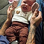 baby, infant, hands, tattoos, clothing, brown_pants, white_shirt, love_text, couch, person, indoor, cute, playful, face, tongue_out, sitting, holding, relaxed, cozy, home