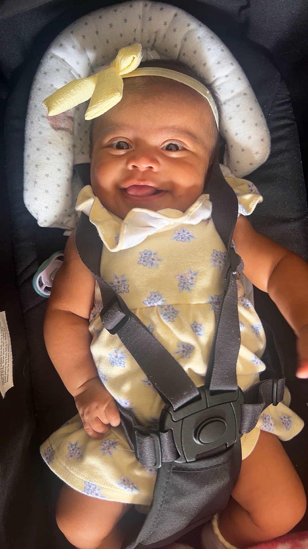 Angie joined the competition — help win amazing prizes! baby, child, smile, car_seat, seatbelt, headband, yellow_dress, floral_pattern, infant, happy, cute, tongue_out, sitting, strapped_in, indoors, closeup, face, skin, hand, footwear