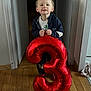 balloon, birthday, boy, casual_clothing, celebration, child, face, happy, holding, home, indoor, light, number_three, party, red, smile, smiling, standing, wooden_floor, young_child