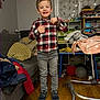 blanket, bookshelf, boy, child, clothing, couch, furniture, happy, indoor, jeans, living_room, person, pillows, plaid_shirt, radiator, smiling, sneakers, standing, toys, wooden_floor
