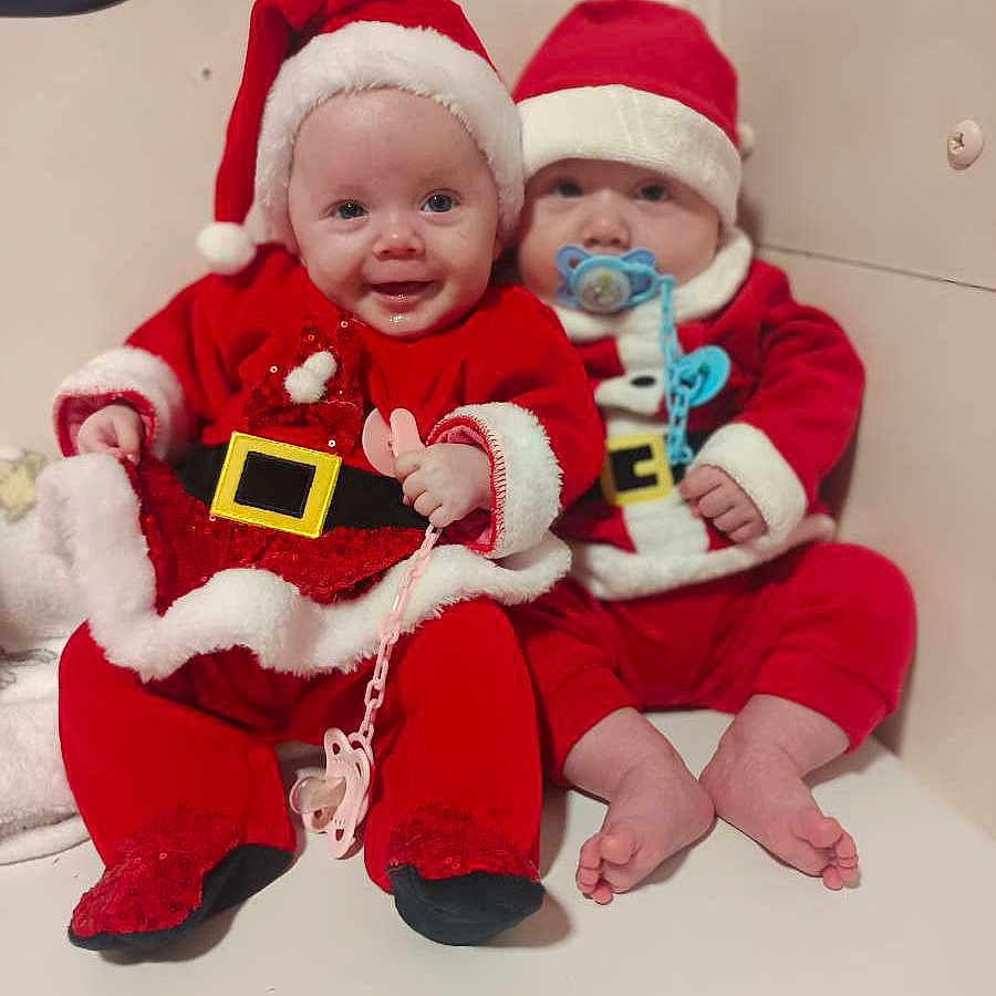 Romy Et Dany a rejoint le concours — aidez-le/la à gagner de superbes lots ! baby, child, christmas, christmas_costume, cute, face, festive, footwear, happy, headwear, holiday, holiday_outfit, indoors, infant, pacifier, red_clothing, santa_hat, seated, smiling, two_babies