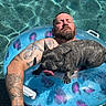 man, dog, french_bulldog, pool, inflatable_ring, tattoo, relaxing, water, sunny, pet, swimming, summer, outdoor, panting, beard, closed_eyes, floating, blue, fun, leisure