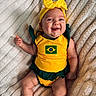baby, infant, smiling, yellow_clothing, headband, bow, blanket, lying_down, cute, happy, face, tongue_out, green_clothing, child, portrait, cheerful, soft_texture, indoors, one_person, skin