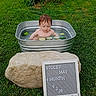 baby, metal_tub, water, cucumber_slices, grass, outdoor, rock, sign, milestone, child, nature, greenery, summer, cute, portrait, infant, sitting, fresh, playful, peaceful