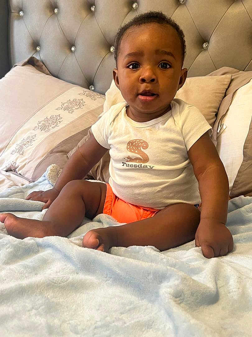 Liam joined the competition — help win amazing prizes! baby, child, toddler, bed, blanket, pillow, white_tshirt, orange_shorts, indoor, home, cute, sitting, portrait, face, skin, head, expression, furniture, cozy, soft