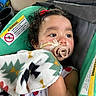 child, baby, pacifier, blanket, car_seat, curly_hair, medical_tape, car_interior, seatbelt, infant, resting, toddler, cute, indoors, closeup, person, seat, warning_label, blanket_pattern, looking_away